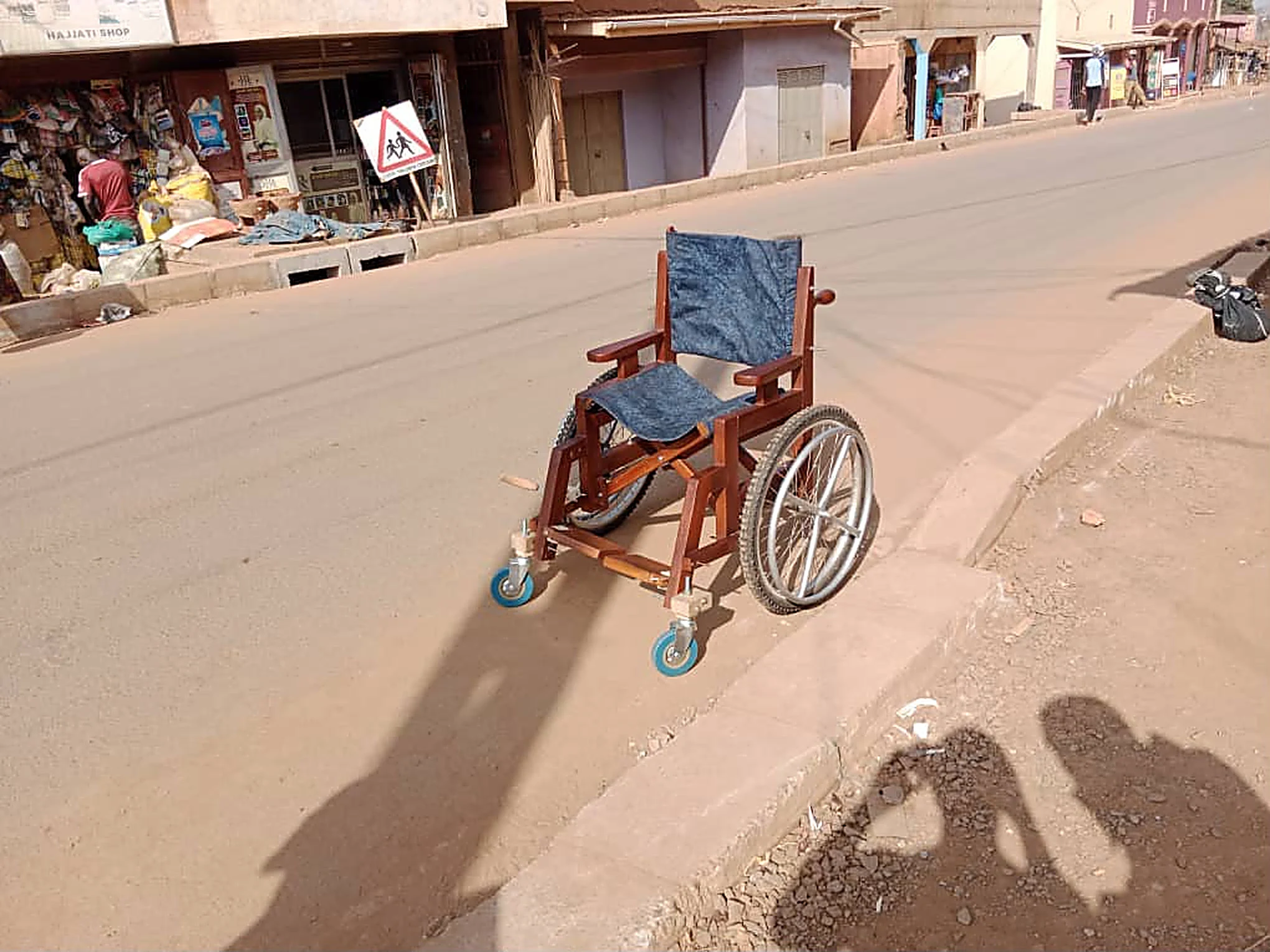 A finished wooden wheelchair with denim seat parked on a red dirt street beside shops.