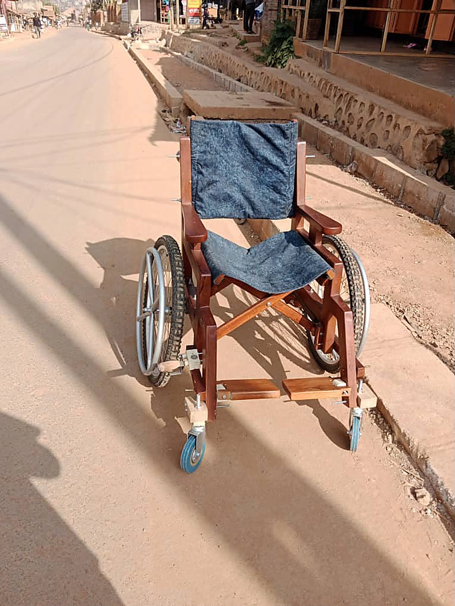 A handcrafted wooden wheelchair with a denim seat, standing on a paved road in Busoga.