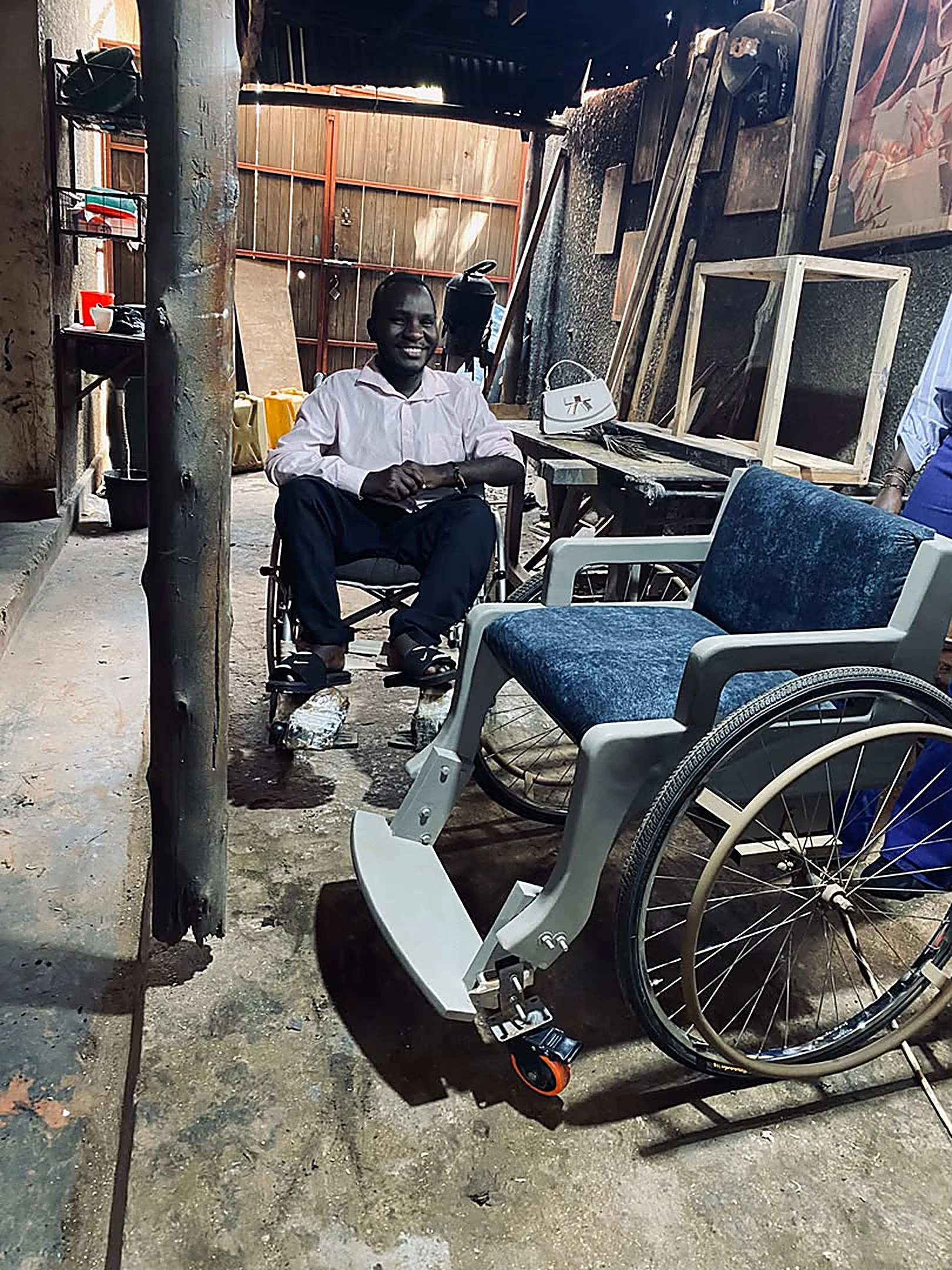 Rashid seated in a wheelchair beside a newly finished terrain wheelchair in the workshop.